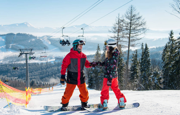 Пара сноубордистов на вершине горнолыжного склона зимнего курорта спускается по склону в солнечный день. Мужчина держит женщину за руку на фоне лыжного подъемника и покрытых снегом гор. Буковель
 - Фото, изображение