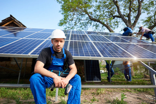 Portrait of successful engineer technician with electrical screwdriver looking in camera, sitting in front of almost finished solar panel photo voltaic system with team of workers on high platform. - Photo, Image