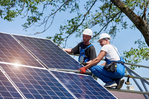Two young technicians mounting heavy solar photo voltaic panel on tall steel platform on green tree background. Exterior solar panel voltaic system installation, dangerous job concept. - 写真・画像