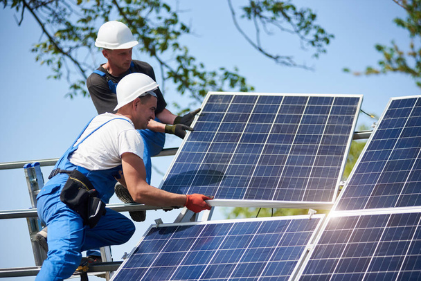 Two workers mounting heavy solar photo voltaic panel on tall steel platform on blue sky background. Exterior stand-alone solar panel system installation, dangerous job concept. - Photo, Image