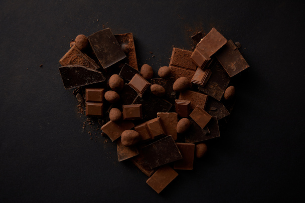 top view of sweet gourmet chocolate pieces with hazelnuts arranged in shape of heart on black - Zdjęcie, obraz