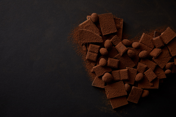bovenaanzicht van heerlijke chocolade stukken met noten en cacao poeder gerangschikt in de vorm van hart op zwarte achtergrond - Foto, afbeelding