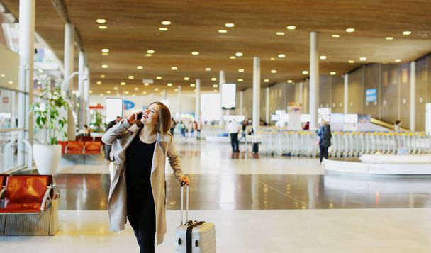 Femme caucasienne avec valise parlant par smartphone dans la salle d'attente de l'aéroport, espace de copie
. - Photo, image