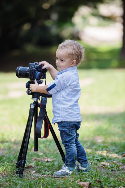 Two years old boy is photographer. Kid holds a camera on tripod and takes photo of landscape. Little child dressed shirt and jeans playing in the park. - Foto, immagini