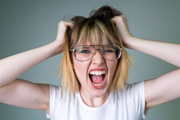 Эмоциональная нервная девушка с светлыми волосами в прозрачных очках, держащая руки за голову, делая гримасу
. - Фото, изображение
