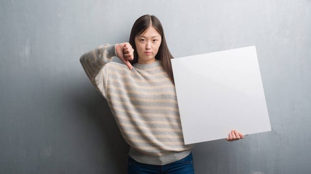 Jeune femme chinoise sur le mur gris tenant la bannière avec le visage en colère, signe négatif montrant aversion avec les pouces vers le bas, concept de rejet
 - Photo, image