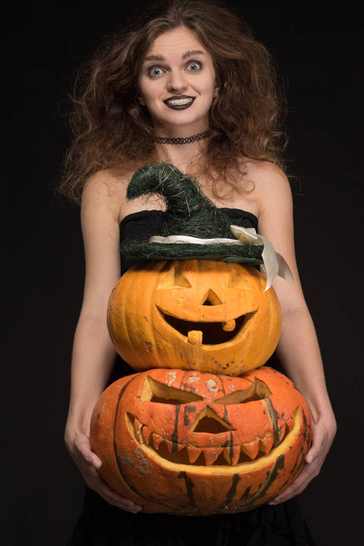 Hermosa chica con un maquillaje gótico como una bruja sonríe y sostiene una calabaza naranja sobre un fondo negro para la fiesta de Halloween
 - Foto, Imagen