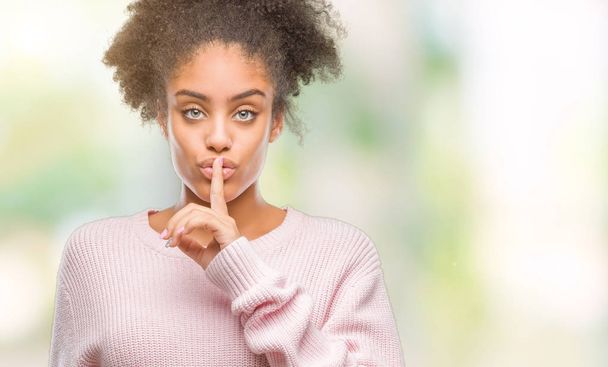 Mujer afroamericana joven que usa suéter de invierno sobre un fondo aislado pidiendo estar callada con el dedo en los labios. Silencio y concepto secreto
. - Foto, Imagen