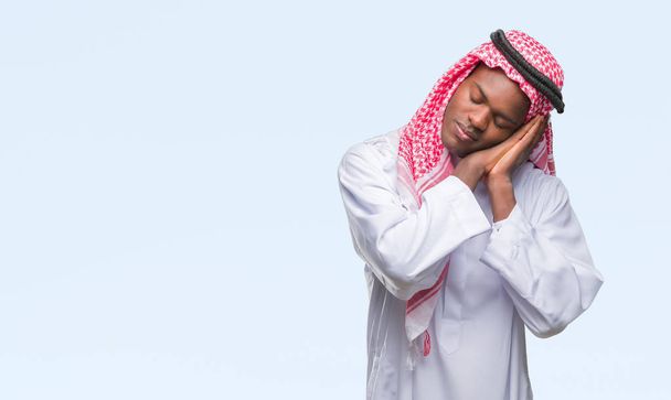Молодой арабический африканский мужчина, одетый в традиционную кеффию на изолированном фоне, спит уставшим от сна и позирует руками, улыбаясь с закрытыми глазами.
. - Фото, изображение