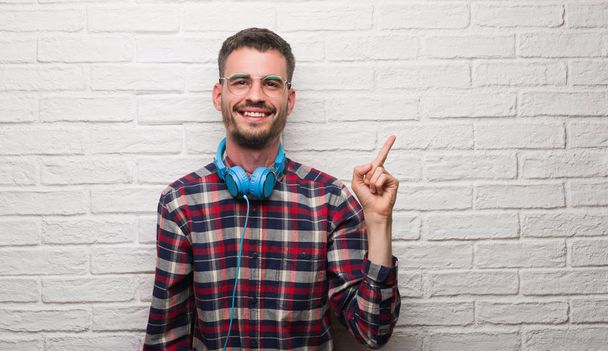 Νεαρός ενήλικος πάνω από τοίχο από τούβλα φορώντας ακουστικά πολύ χαρούμενος, δείχνοντας με το χέρι και δάχτυλο προς την πλευρά - Φωτογραφία, εικόνα
