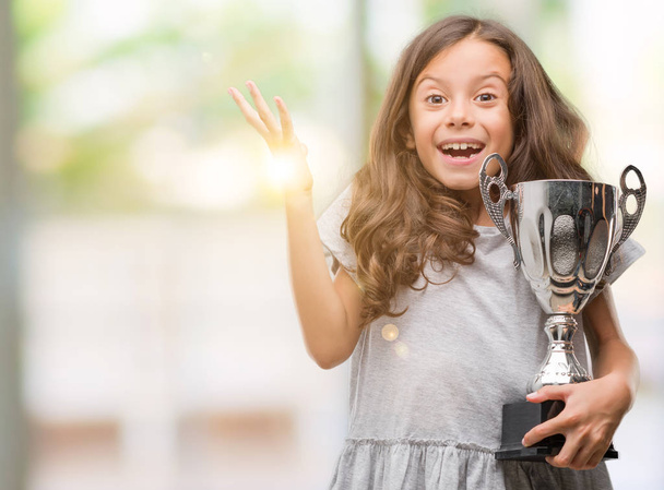 Brunette hispanic girl holding a trophy very happy and excited, winner expression celebrating victory screaming with big smile and raised hands - Foto, afbeelding