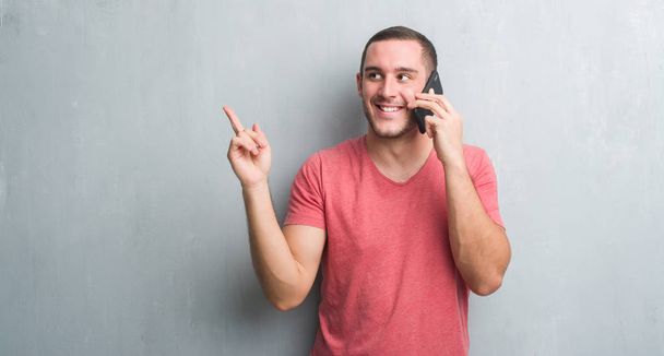 Νεαρός Καυκάσιος πάνω από τοίχο γκρι grunge μιλώντας στο τηλέφωνο πολύ χαρούμενος, δείχνοντας με το χέρι και δάχτυλο προς την πλευρά - Φωτογραφία, εικόνα
