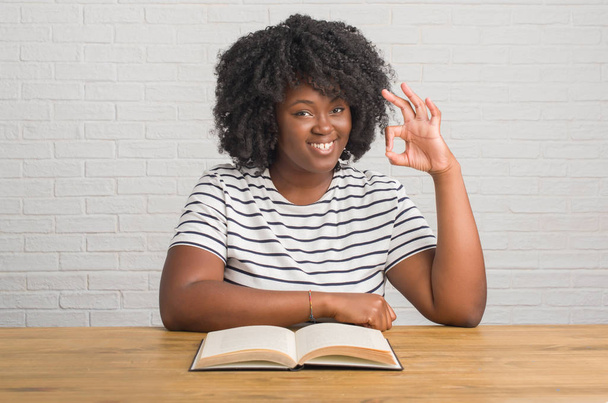 junge afrikanisch-amerikanische Frau sitzt auf dem Tisch und liest Buch und tut ok Zeichen mit den Fingern, ausgezeichnetes Symbol - Foto, Bild
