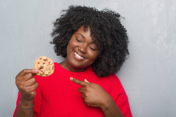 Молода жінка над сірий гранж стіни їсть шоколадної стружки печиво дуже щасливі, вказуючи з рук і пальцями - Фото, зображення