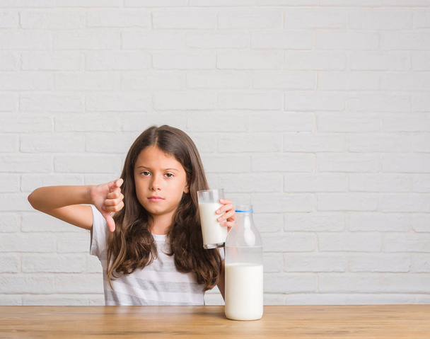 Fiatal spanyol gyerek ül az asztalon, egy pohár tej, haragos arc, negatív előjellel mutatja ellenszenv a hüvelykujját lefelé elutasítás koncepció - Fotó, kép