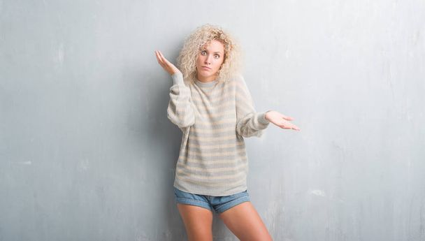 junge blonde Frau mit lockigem Haar über grunge-grauem Hintergrund ahnungslos und verwirrt mit erhobenen Armen und Händen. Zweifelhaftes Konzept. - Foto, Bild