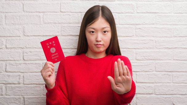 Fiatal kínai nő, nyitott kéz, ezzel súlyos és magabiztos kifejezés, védelmi gesztus stoptábla japán útlevéllel tégla falon - Fotó, kép