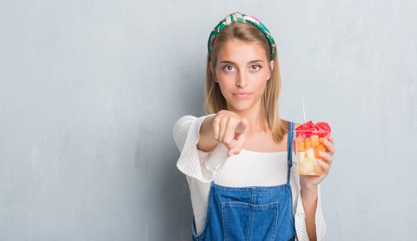 Belle jeune femme sur le mur gris grunge mangeant des fruits pointant du doigt vers la caméra et vers vous, signe de la main, geste positif et confiant de l'avant
 - Photo, image