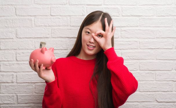 Jonge Chinese vrouw over bakstenen muur piggy bank holding met blij gezicht lachend doen ok teken met de hand op oog kijken door de vingers - Foto, afbeelding