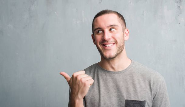 Jeune homme caucasien sur le mur gris grunge pointant et montrant le pouce sur le côté avec le visage heureux souriant
 - Photo, image