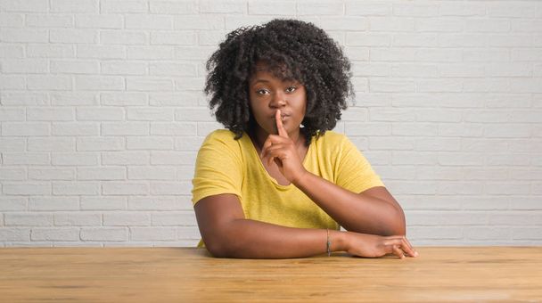 Nuori afrikkalainen amerikkalainen nainen istuu pöydällä kotona pyytäen olla hiljaa sormi huulilla
 - Valokuva, kuva
