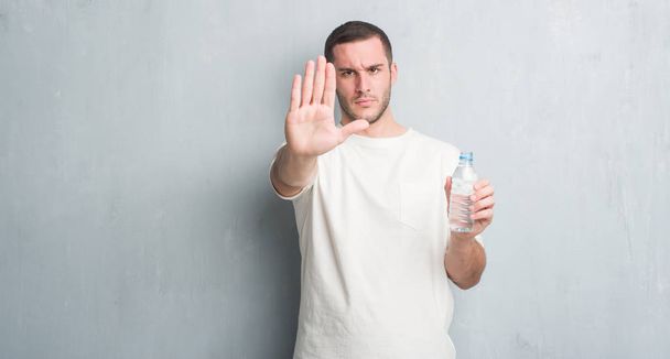 水のボトルを保持している灰色のグランジの壁を越えて若い白人男が防衛ジェスチャー、深刻な自信を持って式に一時停止の標識をやって手を開いてください。 - 写真・画像