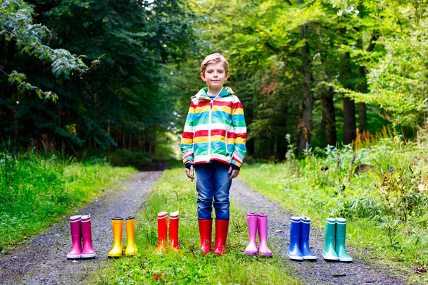Μικρό παιδί αγόρι και ομάδα από πολύχρωμα βροχή μπότες. Ξανθός παιδί στέκεται σε Φθινοπωρινό δάσος. Γκρο πλαν του schoolkid και διαφορετική λαστιχένιες μπότες. Υποδημάτων και αξεσουάρ μόδας για το βροχερό φθινόπωρο - Φωτογραφία, εικόνα