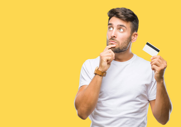 Young handsome man holding credit card over isolated background serious face thinking about question, very confused idea - Фото, изображение