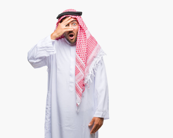 Όμορφος νεαρός φορώντας keffiyeh πέρα από το απομονωμένο υπόβαθρο peeking σε κατάσταση σοκ που καλύπτουν το πρόσωπο και τα μάτια με το χέρι, κοιτάζοντας μέσα από τα δάχτυλα με αμηχανία έκφρασης. - Φωτογραφία, εικόνα