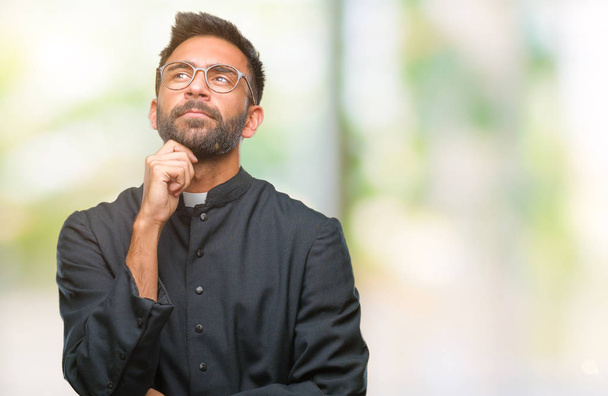 Erwachsener hispanischer katholischer Priester über isoliertem Hintergrund mit der Hand am Kinn, der über Frage nachdenkt, nachdenklichen Ausdruck. Lächeln mit nachdenklichem Gesicht. Zweifelhaftes Konzept. - Foto, Bild