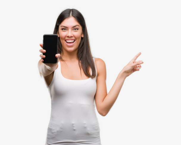 Young beautiful hispanic showing smartphone very happy pointing with hand and finger to the side - Foto, imagen