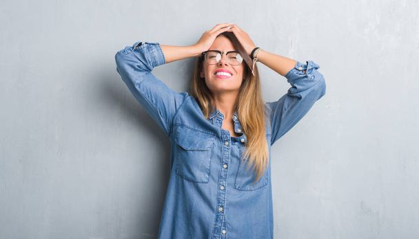 Mujer adulta joven sobre la pared gris grunge usando gafas que sufren de dolor de cabeza desesperada y estresada porque el dolor y la migraña. Manos en la cabeza
. - Foto, Imagen