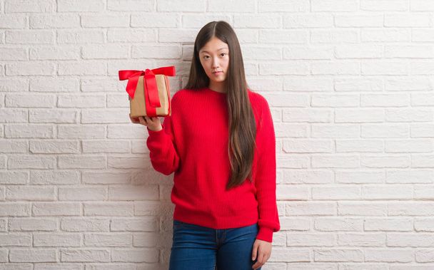 Молодая китаянка за кирпичной стеной держит подарок с уверенным выражением на умном лице, думая серьезно
 - Фото, изображение