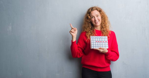 junge rothaarige Frau über graue Grunge-Wand mit Zeitrahmen-Kalender sehr glücklich mit Hand und Finger zur Seite zeigend - Foto, Bild