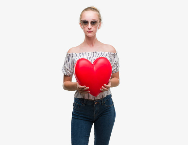 Blonde teenager woman holding red heart with a confident expression on smart face thinking serious - Foto, afbeelding