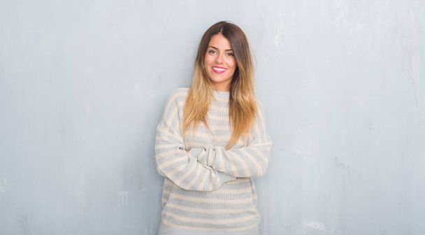 Молодая взрослая женщина над гранж-серой стеной в зимнем свитере счастливое лицо улыбается со скрещенными руками, глядя в камеру. Позитивный человек
. - Фото, изображение