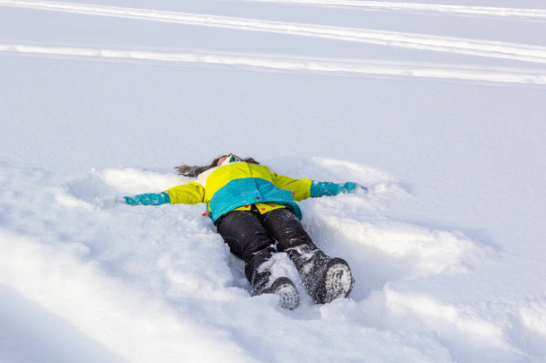 Chica linda sonriente alegre y feliz tumbado en la nieve y haciendo ángel de nieve, aleteando sus brazos y piernas en diferentes direcciones, disfrutando y regocijándose invierno
. - Foto, Imagen