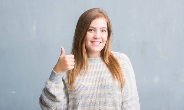 Молодая взрослая женщина над серой гранж-стеной счастлива с большой улыбкой делает хорошо знак, палец вверх пальцами, отличный знак
 - Фото, изображение