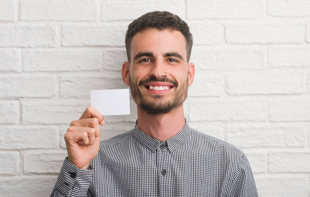 Joven hombre adulto sobre pared de ladrillo sosteniendo la tarjeta en blanco con una cara feliz de pie y sonriendo con una sonrisa confiada mostrando los dientes
 - Foto, imagen