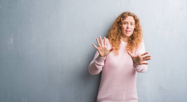 Jonge roodharige vrouw over grijs grunge muur dragen roze trui bang en doodsbang met angst expressie stop gebaar met handen, schreeuwen in een roes. Paniek concept. - Foto, afbeelding