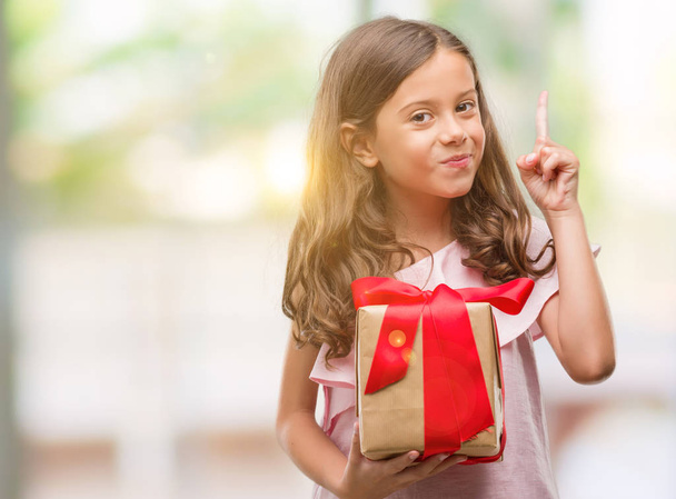 Brunetka hispánský dívka držící dárek překvapilo s nápad nebo otázku ukazující prst s šťastné tváře, číslo jedna - Fotografie, Obrázek