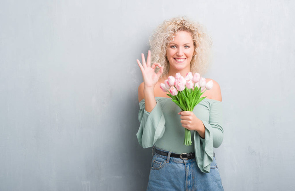 Young blonde woman over grunge grey background holding flowers doing ok sign with fingers, excellent symbol - Photo, Image