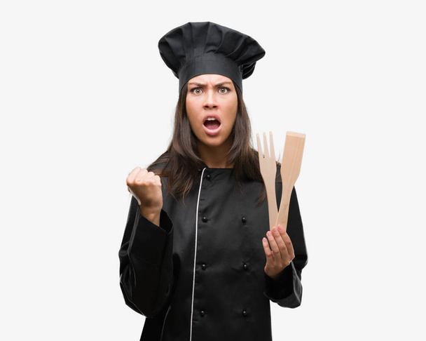 Jeune cuisinière hispanique portant l'uniforme de chef ennuyée et frustrée criant de colère, folle et criant avec la main levée, concept de colère
 - Photo, image