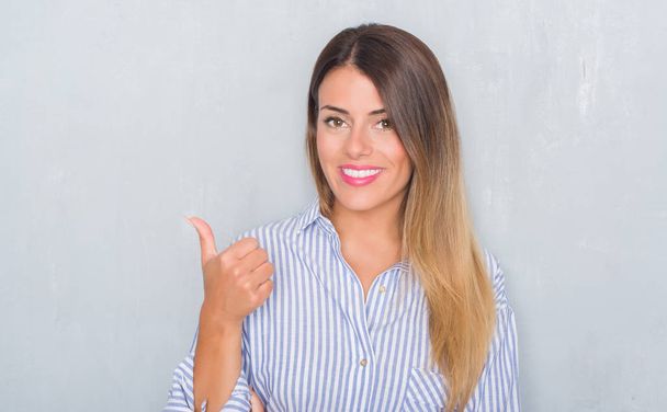 Jonge volwassen vrouw over grunge grijs muur dragen business shirt glimlachend met blij gezicht kijken en naar de kant met duim omhoog. - Foto, afbeelding