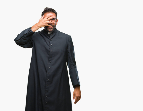 Ενηλίκων Ισπανόφωνος καθολικός ιερέας άνδρα πάνω σε μια απομονωμένη φόντο peeking σε κατάσταση σοκ που καλύπτουν το πρόσωπο και τα μάτια με το χέρι, κοιτάζοντας μέσα από τα δάχτυλα με αμηχανία έκφρασης. - Φωτογραφία, εικόνα