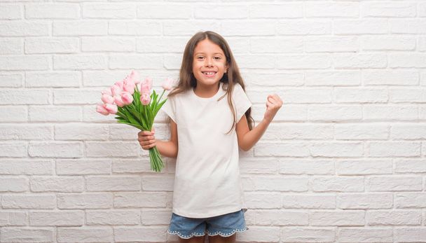 Young hispanic kid over white brick wall holding flowers on mother day screaming proud and celebrating victory and success very excited, cheering emotion - Photo, Image