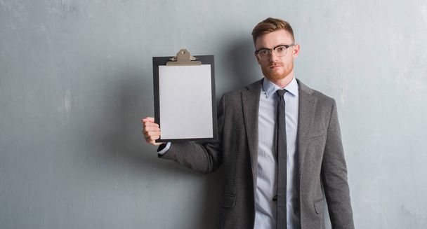Jonge roodharige zakenman over grijs grunge muur holding Klembord met een vertrouwen uitdrukking op slimme gezicht ernstige denken - Foto, afbeelding