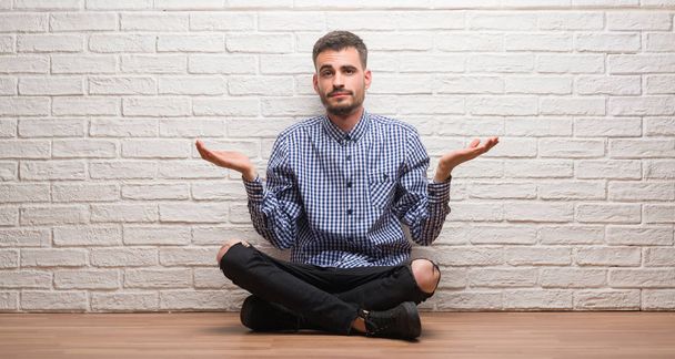 Hombre adulto joven sentado sobre la pared de ladrillo blanco expresión despistado y confundido con los brazos y las manos levantadas. Concepto de duda
. - Foto, imagen