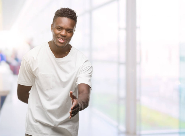 Молодой африканский американец в белой футболке с дружеской улыбкой предлагает рукопожатие в качестве приветствия и приветствия. Успешный бизнес
. - Фото, изображение