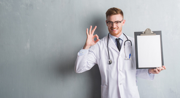Mladá zrzka doktor muž přes zeď šedá grunge drží schránky dělá ok znamení prsty, vynikající symbol - Fotografie, Obrázek
